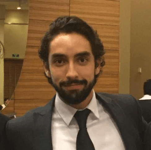 Bearded man in a suit and tie posing indoors with wooden panel background.